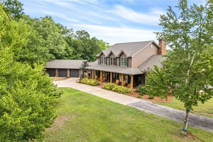 A classic country home, featuring a generous front porch and attached three-car garage, is nestled among mature trees.