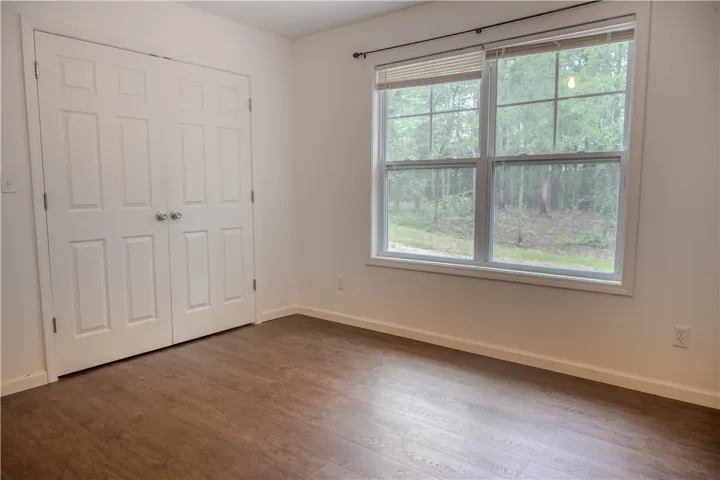 A well-lit bedroom features sturdy wood-look flooring and a generous window overlooking green trees.