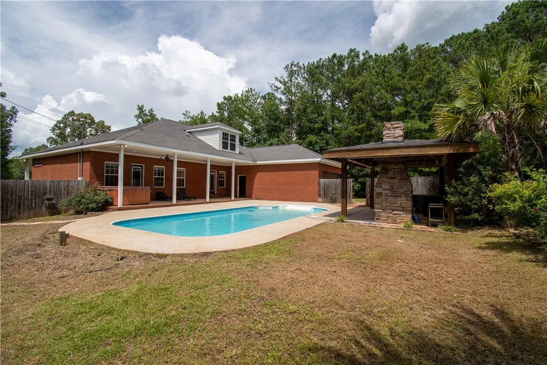 An inviting pool alongside a brick residence, complemented by a covered outdoor living area with a stone fireplace.