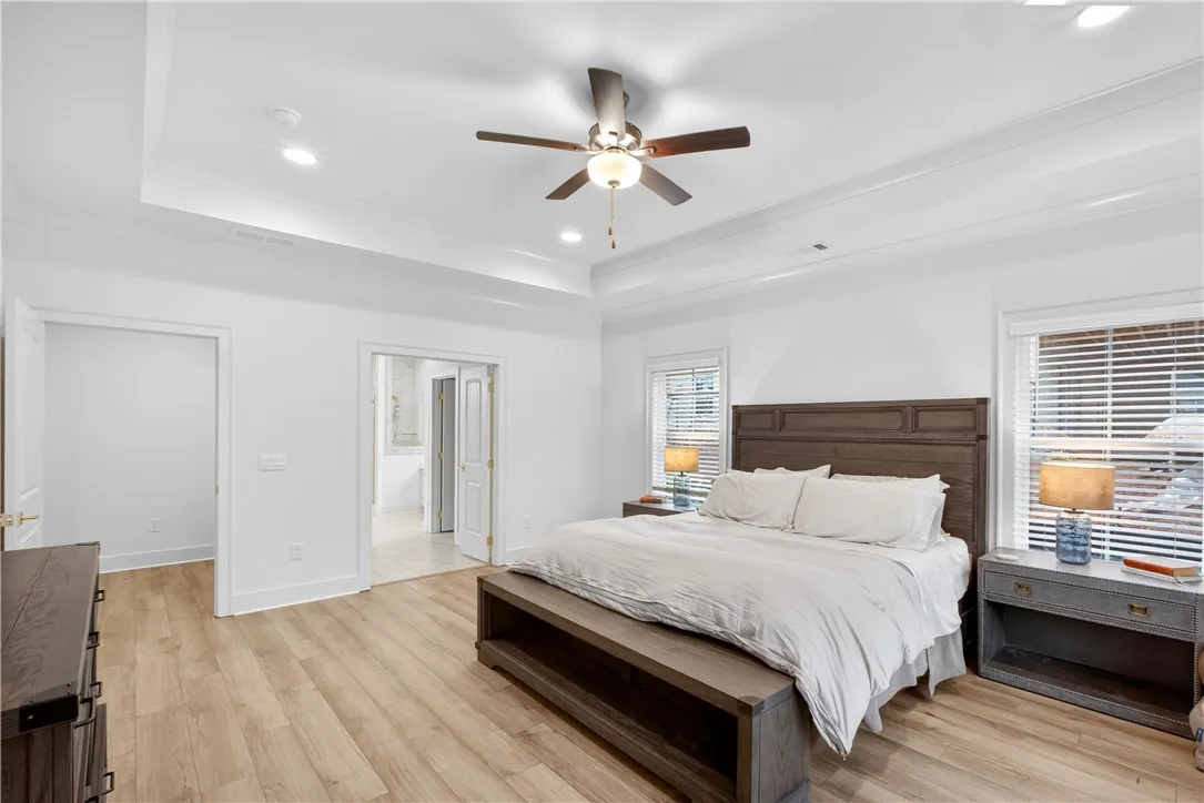 Recessed lighting and a ceiling fan illuminate this spacious bedroom with warm-toned flooring and tray ceilings.