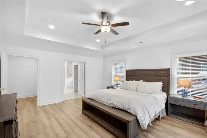 Recessed lighting and a ceiling fan illuminate this spacious bedroom with warm-toned flooring and tray ceilings.
