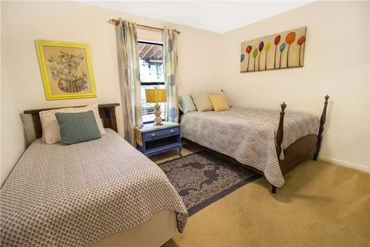 A well-lit guest bedroom featuring two beds, soft carpet flooring, and a window with nature views.