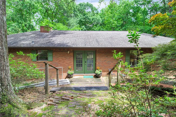 Mature trees canopy this inviting brick home, featuring green French doors and a charming, moss-kissed brick path.