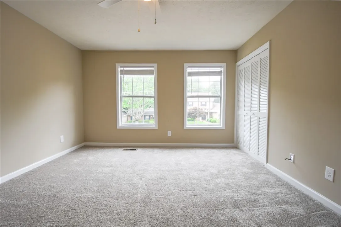 A well-lit bedroom features plush gray carpeting and dual windows overlooking a peaceful neighborhood.