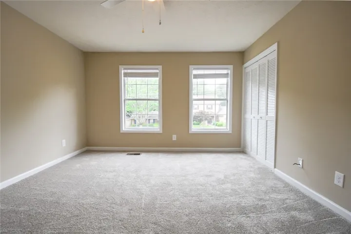 A well-lit bedroom features plush gray carpeting and dual windows overlooking a peaceful neighborhood.
