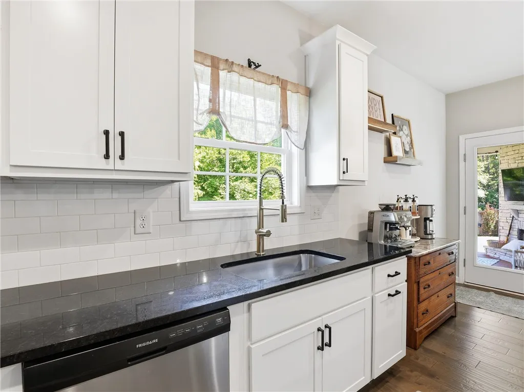 A well-lit kitchen features white cabinetry, dark countertops, and a full-size sink overlooking the verdant outdoors.