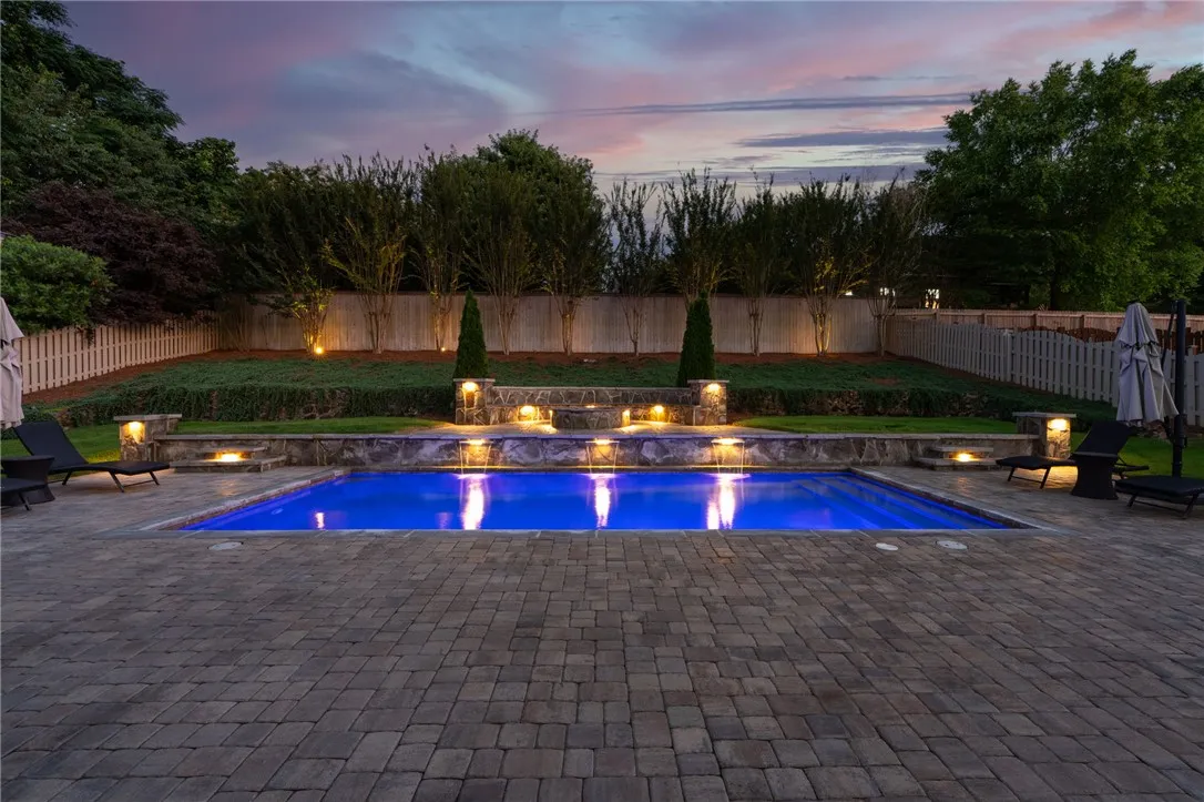 An illuminated backyard oasis with a pristine pool and charming stone waterfall feature, perfect for evening enjoyment.