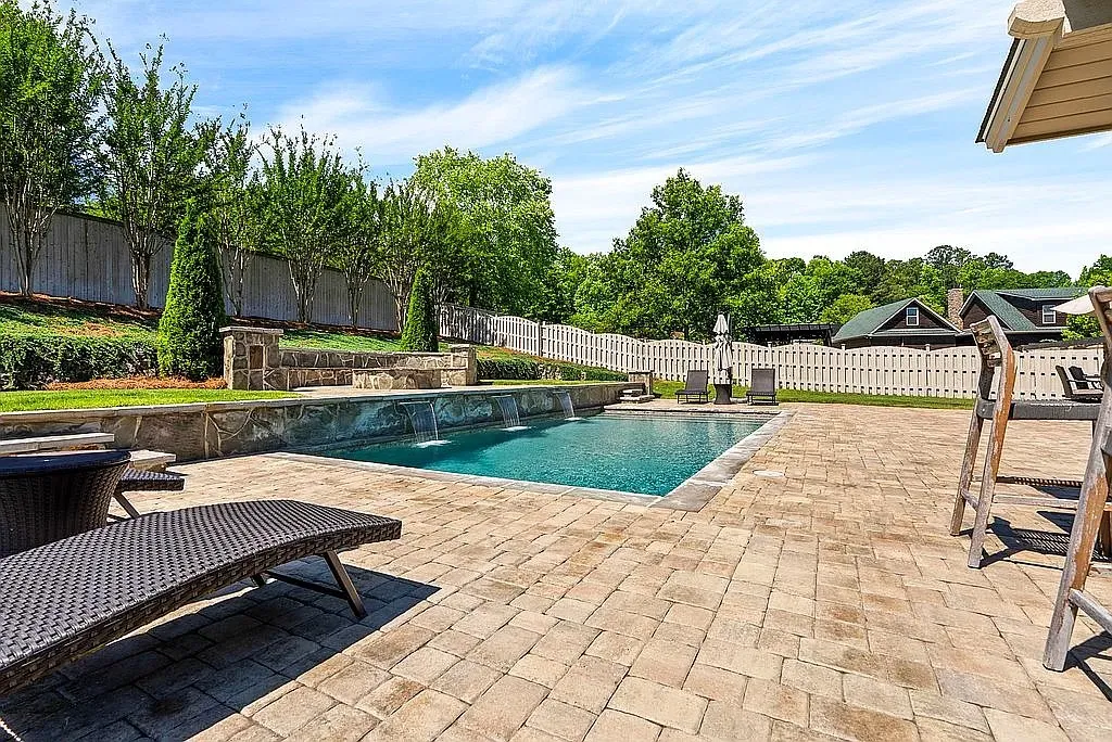 An inviting pool area with cascading water features awaits, surrounded by lush greenery and a spacious paver patio.