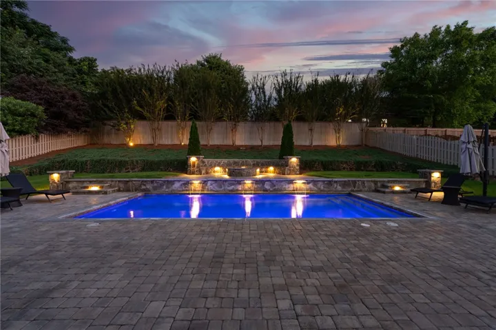 An illuminated backyard oasis with a pristine pool and charming stone waterfall feature, perfect for evening enjoyment.
