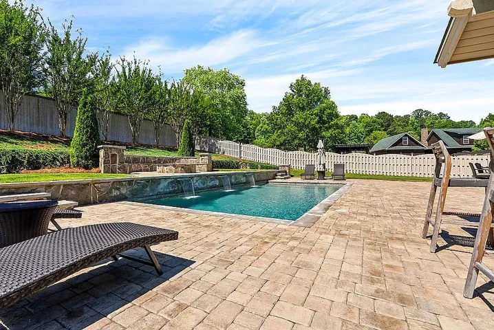 An inviting pool area with cascading water features awaits, surrounded by lush greenery and a spacious paver patio.