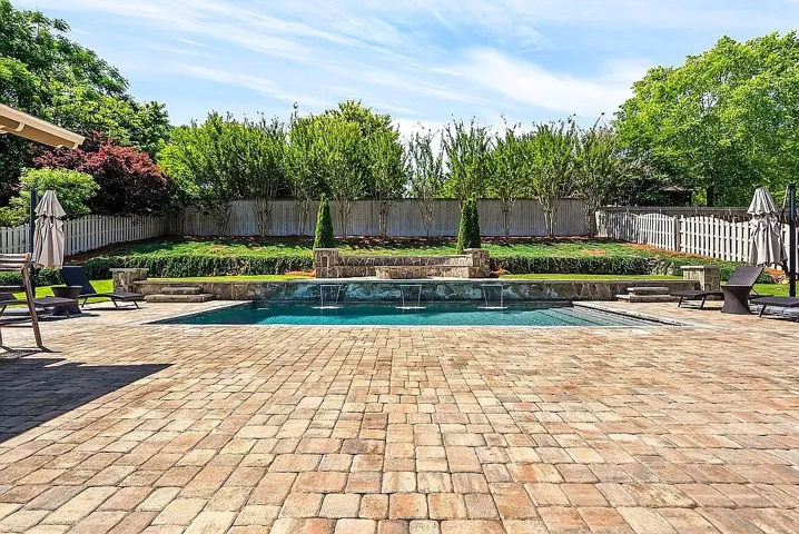 An inviting pool area awaits, framed by lush landscaping and a classic picket fence.