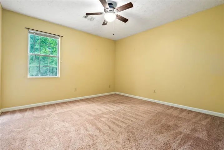 A well-lit bedroom features plush carpeting, a single window, and a ceiling fan for comfort.