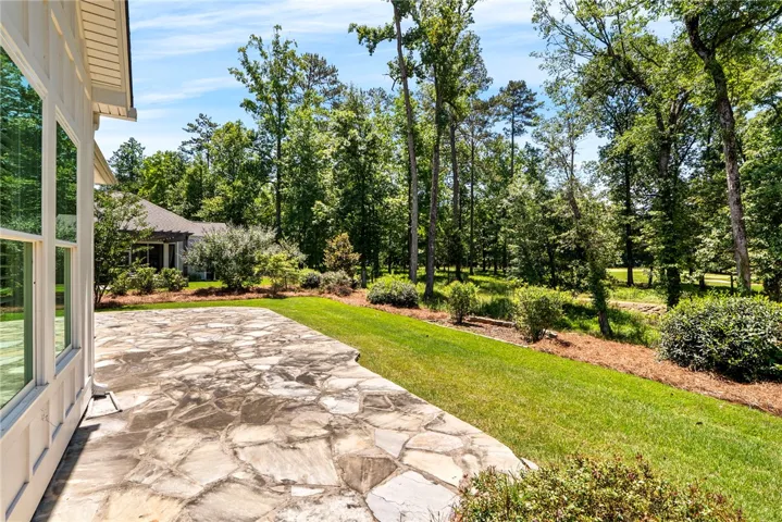 An inviting stone patio unfolds into a verdant landscape, bordered by mature trees and natural plantings.