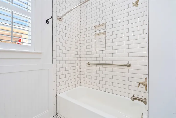 The bathroom features a clean white bathtub and classic subway tile surround, illuminated by natural light from a window with white shutters.