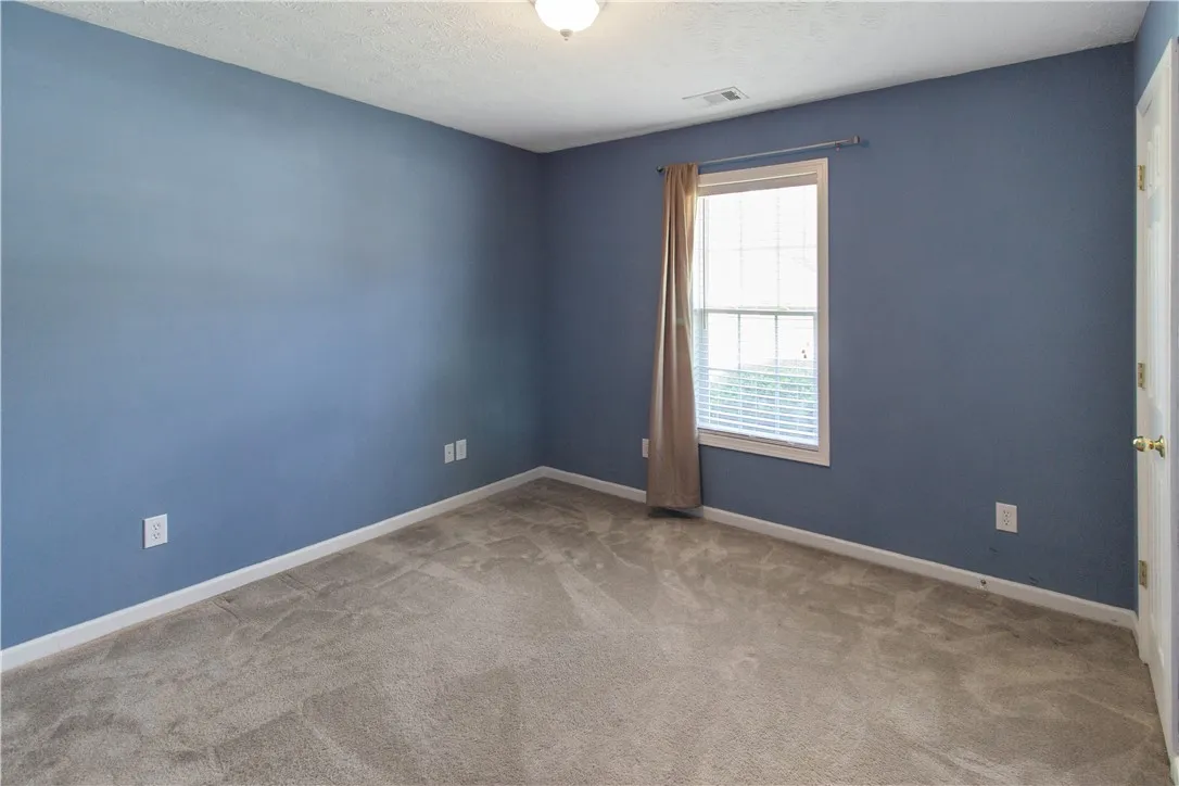 A well-lit bedroom features plush carpeting and a bright window with neutral drapes.