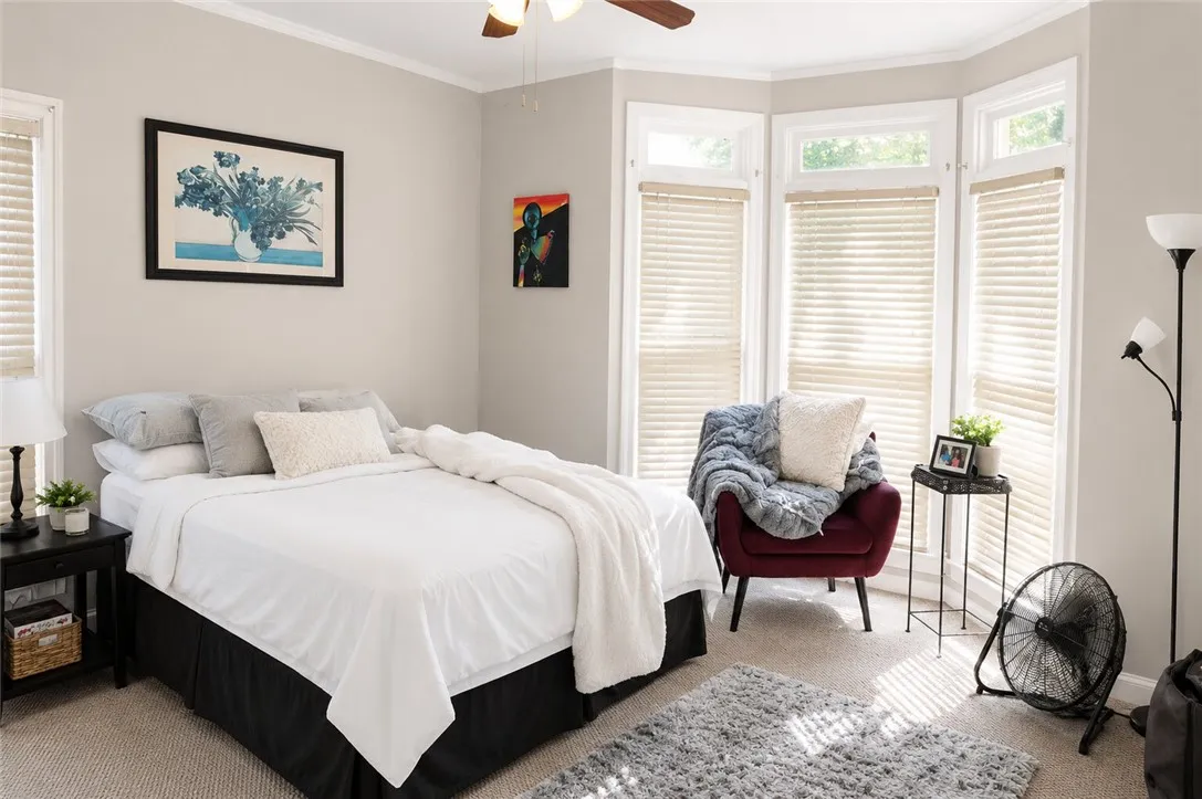 A comfortable bedroom features bay windows, soft carpeting, and a cooling ceiling fan.
