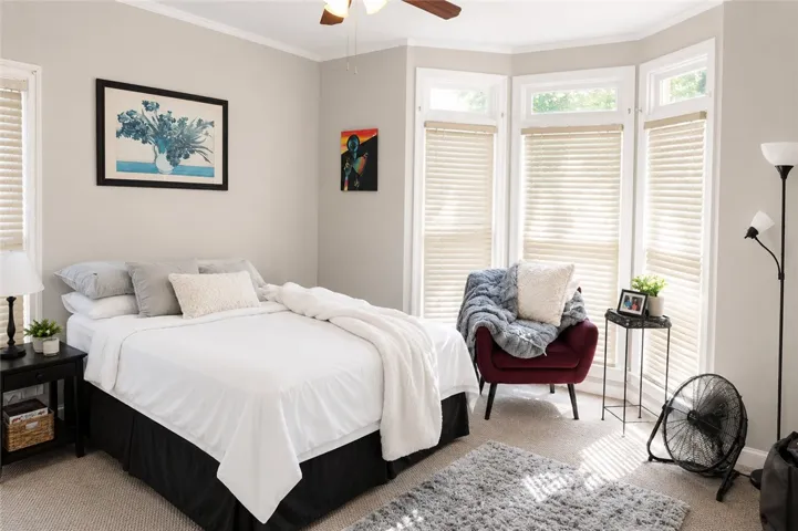 A comfortable bedroom features bay windows, soft carpeting, and a cooling ceiling fan.