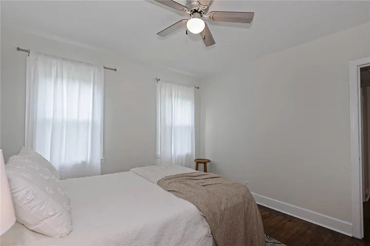 Hardwood flooring grounds this serene bedroom, featuring twin windows draped in sheer curtains and a ceiling fan for optimal comfort.