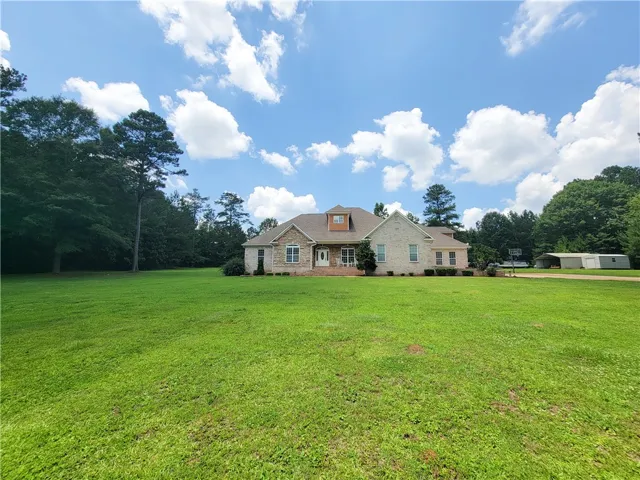 A spacious front yard unfolds before a stately stone and brick home, framed by a serene, wooded backdrop.