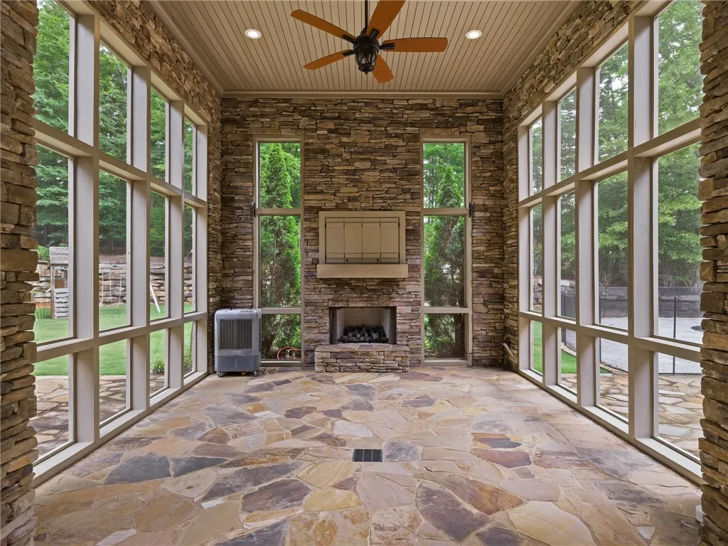 A spacious sunroom with flagstone flooring features a stone fireplace and tall, multi-pane windows overlooking the lush yard.