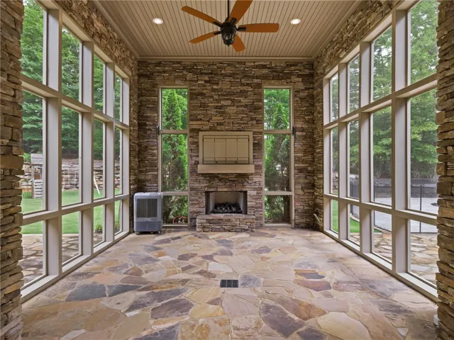 A spacious sunroom with flagstone flooring features a stone fireplace and tall, multi-pane windows overlooking the lush yard.