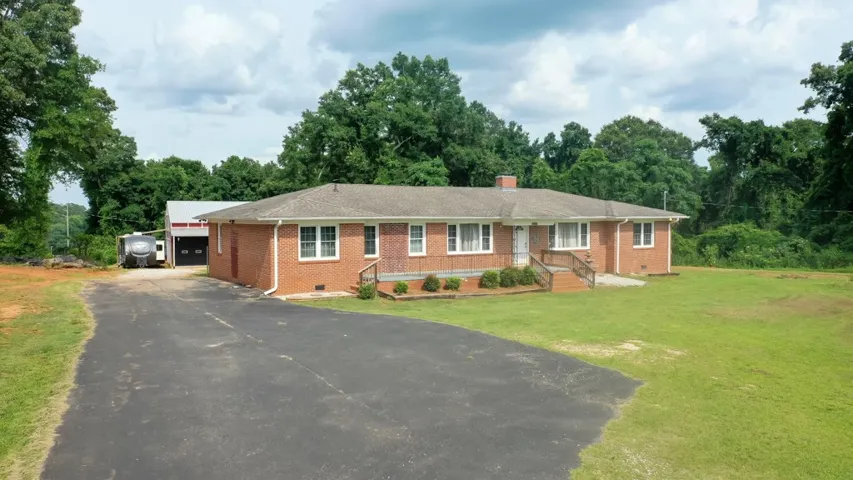 A spacious ranch home with a long asphalt driveway, set against a backdrop of mature trees and a separate workshop.