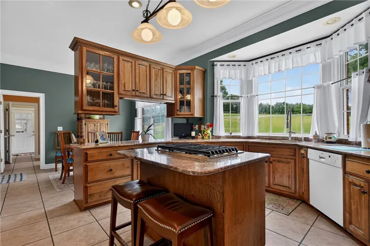 A country-style kitchen features a gas cooktop island and a bay window overlooking vast green fields.