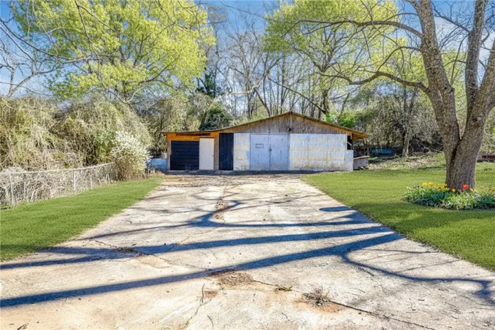A long concrete driveway leads to a large outbuilding, framed by mature trees and lush grass.