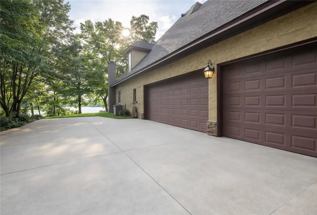 A spacious concrete driveway leads to a two-car garage, framed by verdant trees and a distant lake.