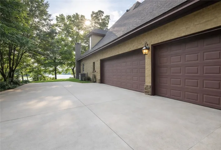 A spacious concrete driveway leads to a two-car garage, framed by verdant trees and a distant lake.