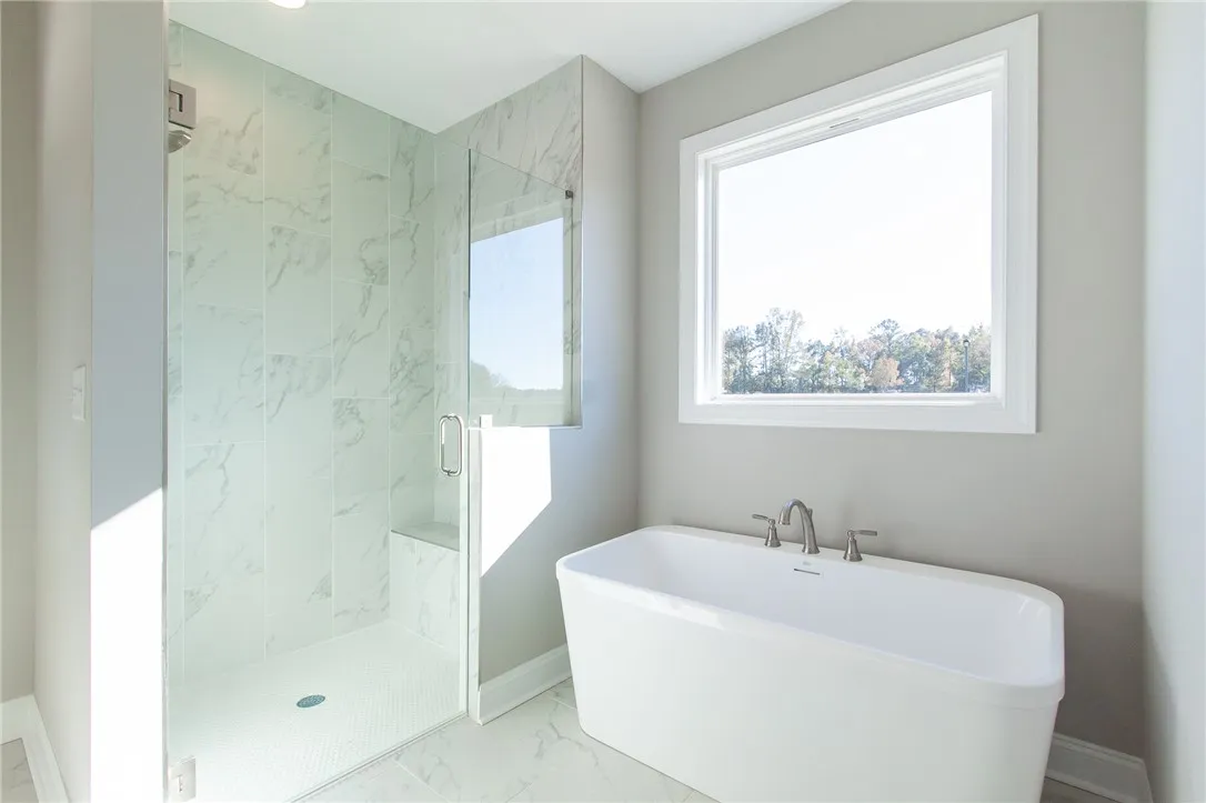 A spacious primary bath features a walk-in shower and a free-standing tub, illuminated by a broad window. Photos are not of subject property. Photos are of the same floorplan of a completed home.