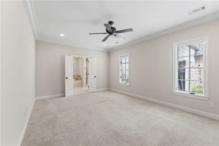 A spacious, naturally illuminated room features plush carpeting and elegant crown molding, with double doors opening to a bath.