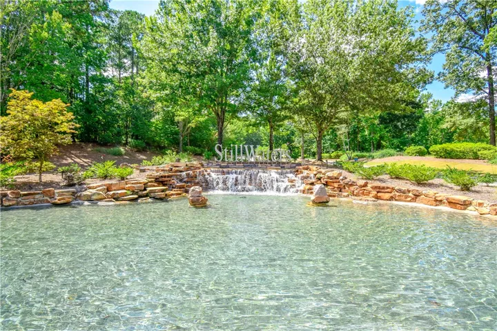 Clear waters flow into a tranquil pond with a stone waterfall feature, nestled amidst lush trees and landscaping.