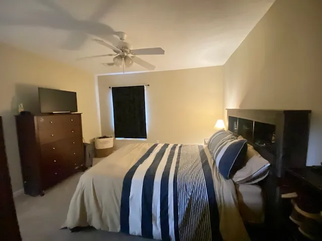 A comfortable bedroom features a ceiling fan, plush carpeting, and a window with black-out shades for peaceful rest.