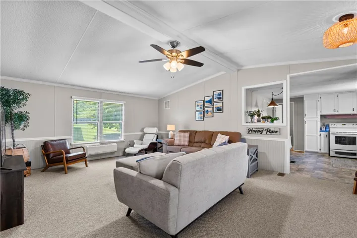 A well-lit living area features a ceiling fan, plush carpeting, and an open pass-through to the kitchen.