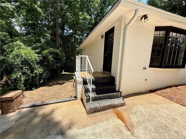 A well-maintained side entrance leads to a charming brick exterior with a large bay window, nestled among mature trees.