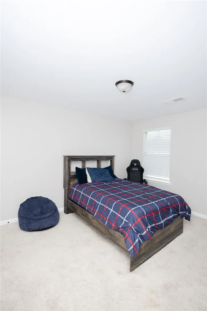 A comfortable bedroom with a window allowing natural light features a textured carpet and a simple ceiling fixture.