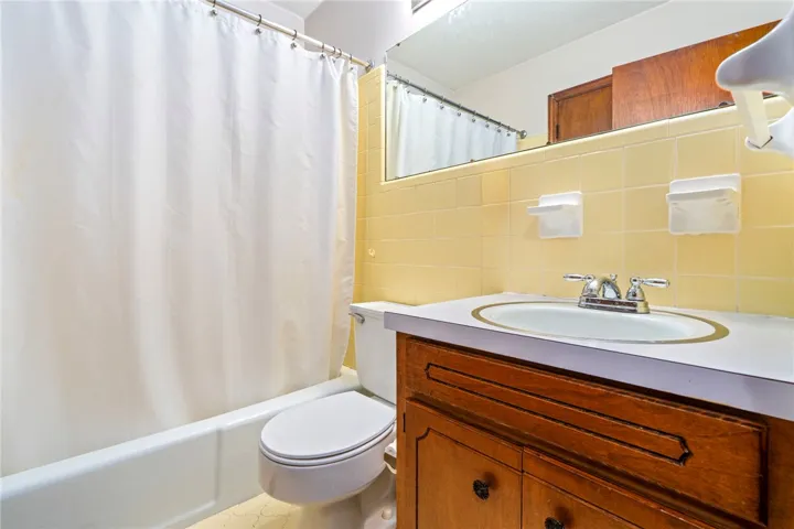 A compact bathroom featuring a full bathtub, classic yellow tiled walls, and a warm wood vanity.