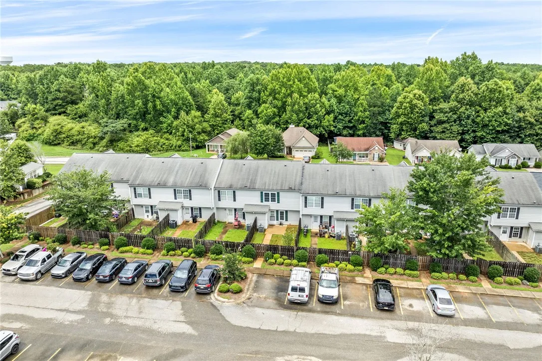 An aerial perspective reveals a row of townhomes with individual fenced yards and ample parking, all set against a backdrop of mature trees.