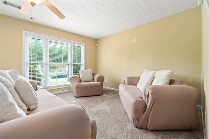 A well-lit living space features a large window offering neighborhood views and soft carpet underfoot.