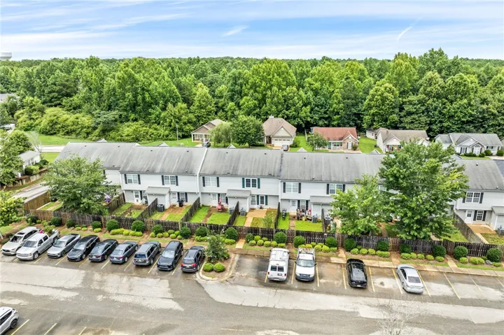 An aerial perspective reveals a row of townhomes with individual fenced yards and ample parking, all set against a backdrop of mature trees.