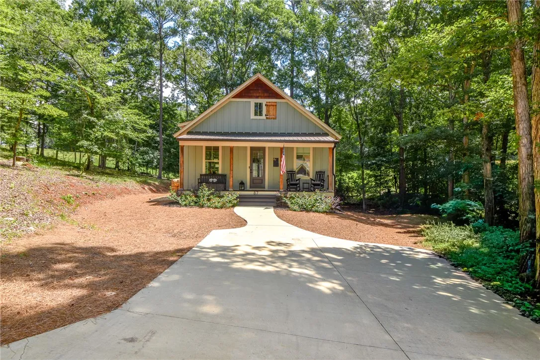 A charming home nestled amongst dense trees, featuring a welcoming covered front porch and a paved driveway.