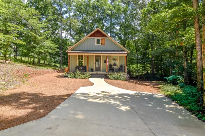 A charming home nestled amongst dense trees, featuring a welcoming covered front porch and a paved driveway.
