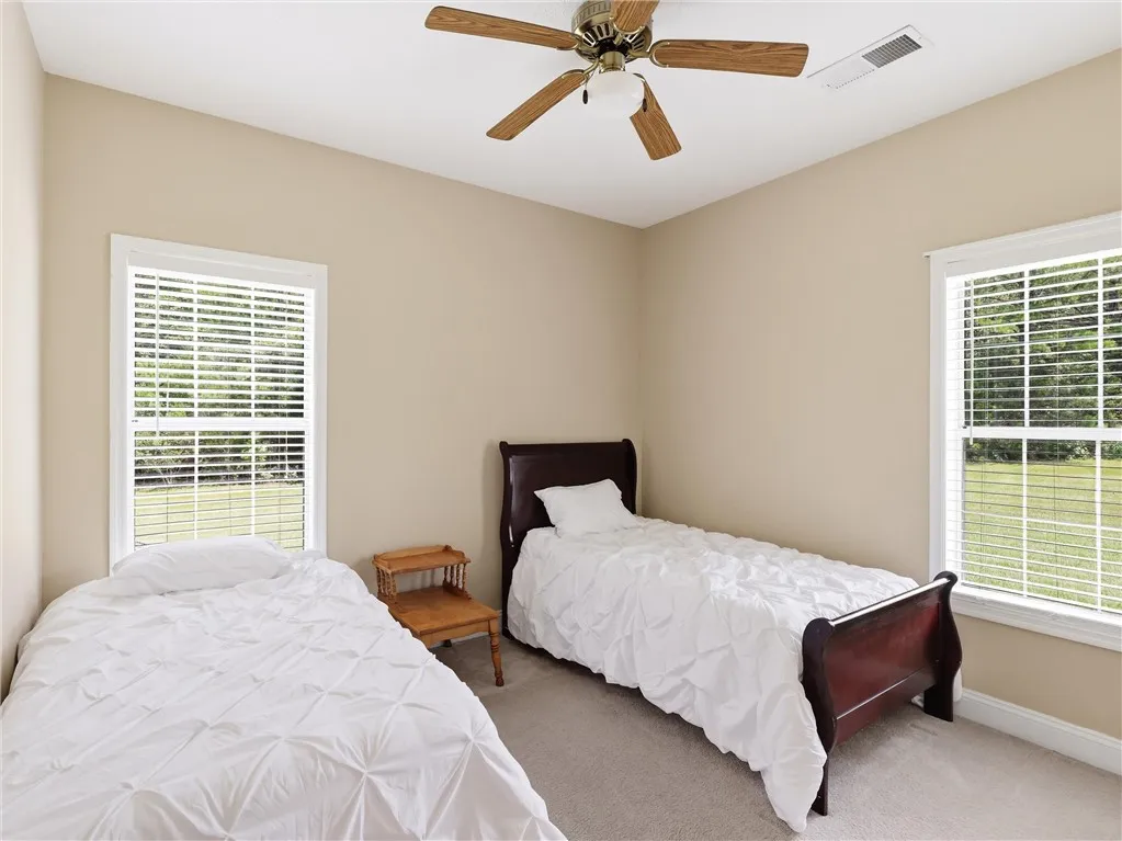 Two bright windows frame a carpeted bedroom, offering views of verdant outdoor greenery.
