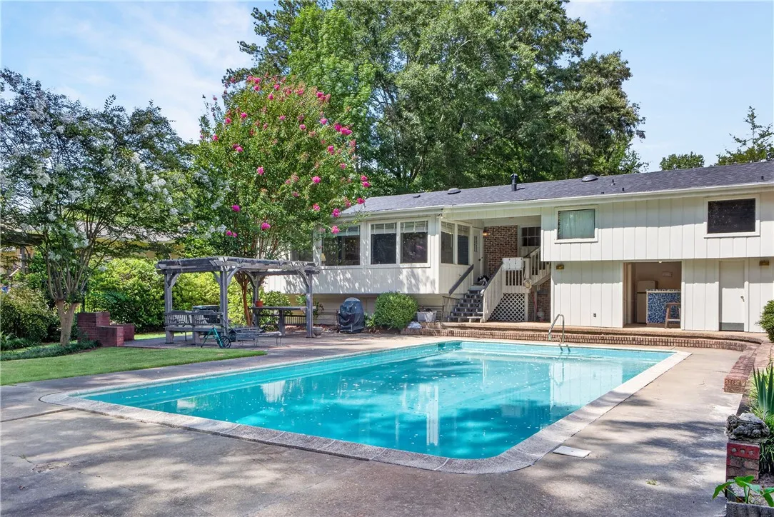 An inviting in-ground pool awaits in a landscaped backyard featuring a shaded pergola and mature trees.