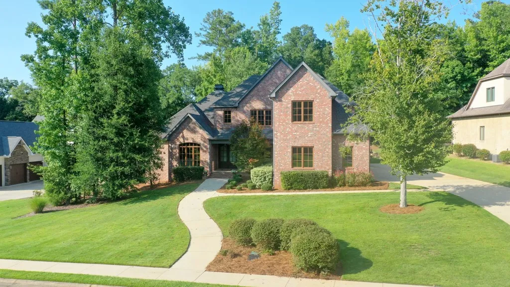 A graceful curved walkway guides visitors to this traditional brick home, set against a backdrop of mature trees and vibrant green landscaping.