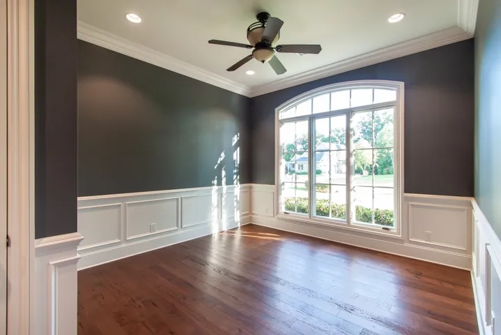 Elegant dark walls frame a sun-drenched bedroom or office with rich wood flooring and classic wainscoting.