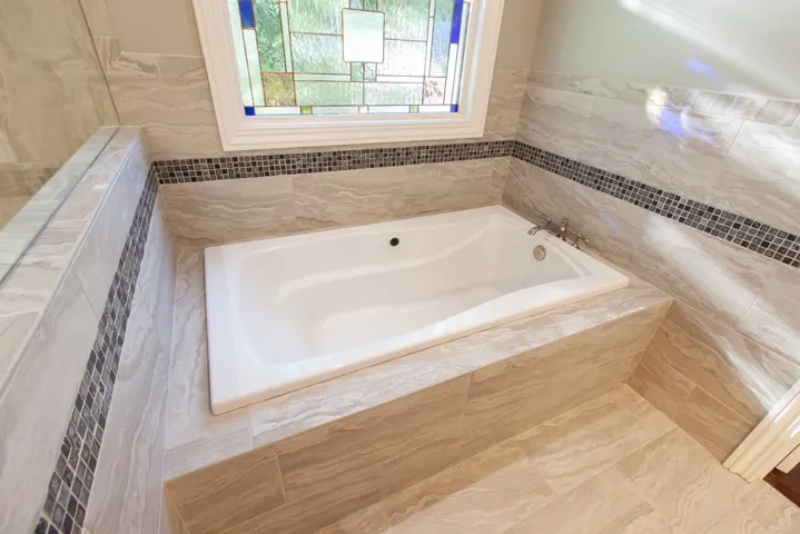 Indulge in a tranquil soak within this well-appointed bath featuring elegant tile work and a charming stained-glass window.