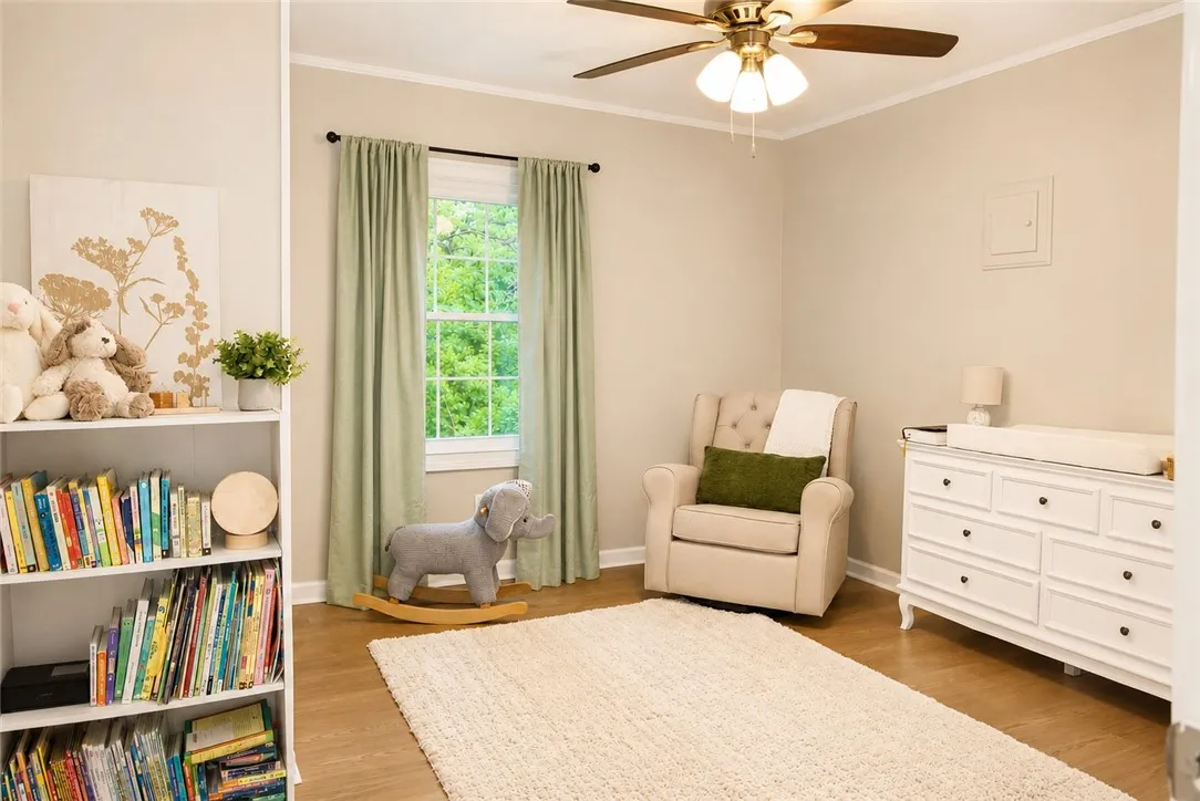 A well-lit nursery features hardwood flooring, a ceiling fan, and a large window with ample natural light.