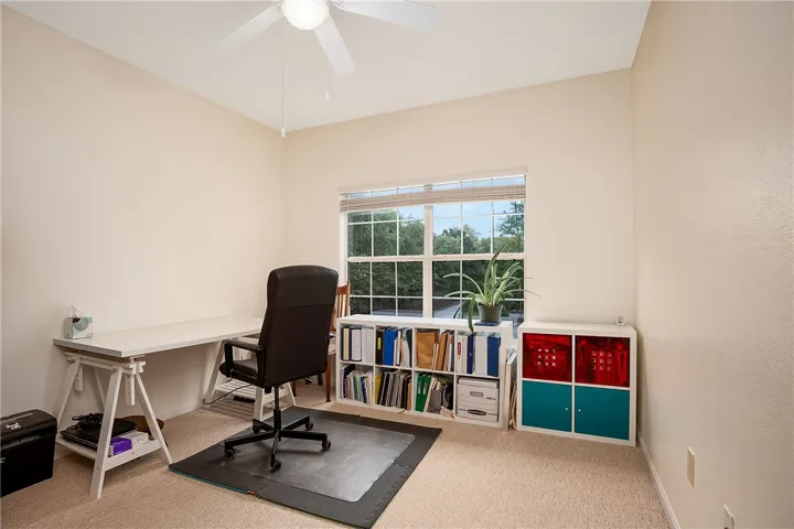A well-lit home office/bedroom features carpeted floors and a prominent window for abundant natural light.